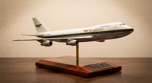 A historic 1:200 diecast scale model of a classic Boeing 747-200 Queen of the Skies in vintage 1970s green cheatline and gold trim livery. Displayed on a solid polished mahogany base with commemorative bronze inscription plate, illuminated by gentle directional museum gallery lighting on a soft off-white canvas.